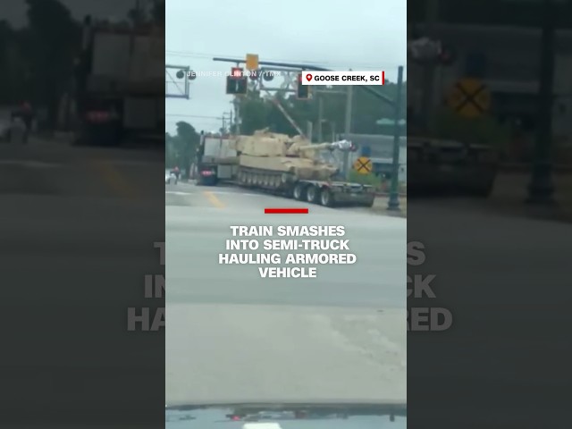 ⁣Drivers watch as a train smashes into a semi-truck hauling an armored vehicle